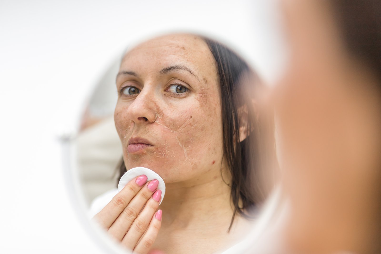 Woman wiping her cheek with a cotton pad, seen in a round mirror