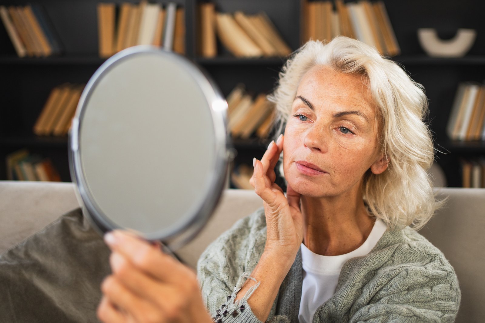 Older woman holding a round hand mirror and examining the skin on her cheek