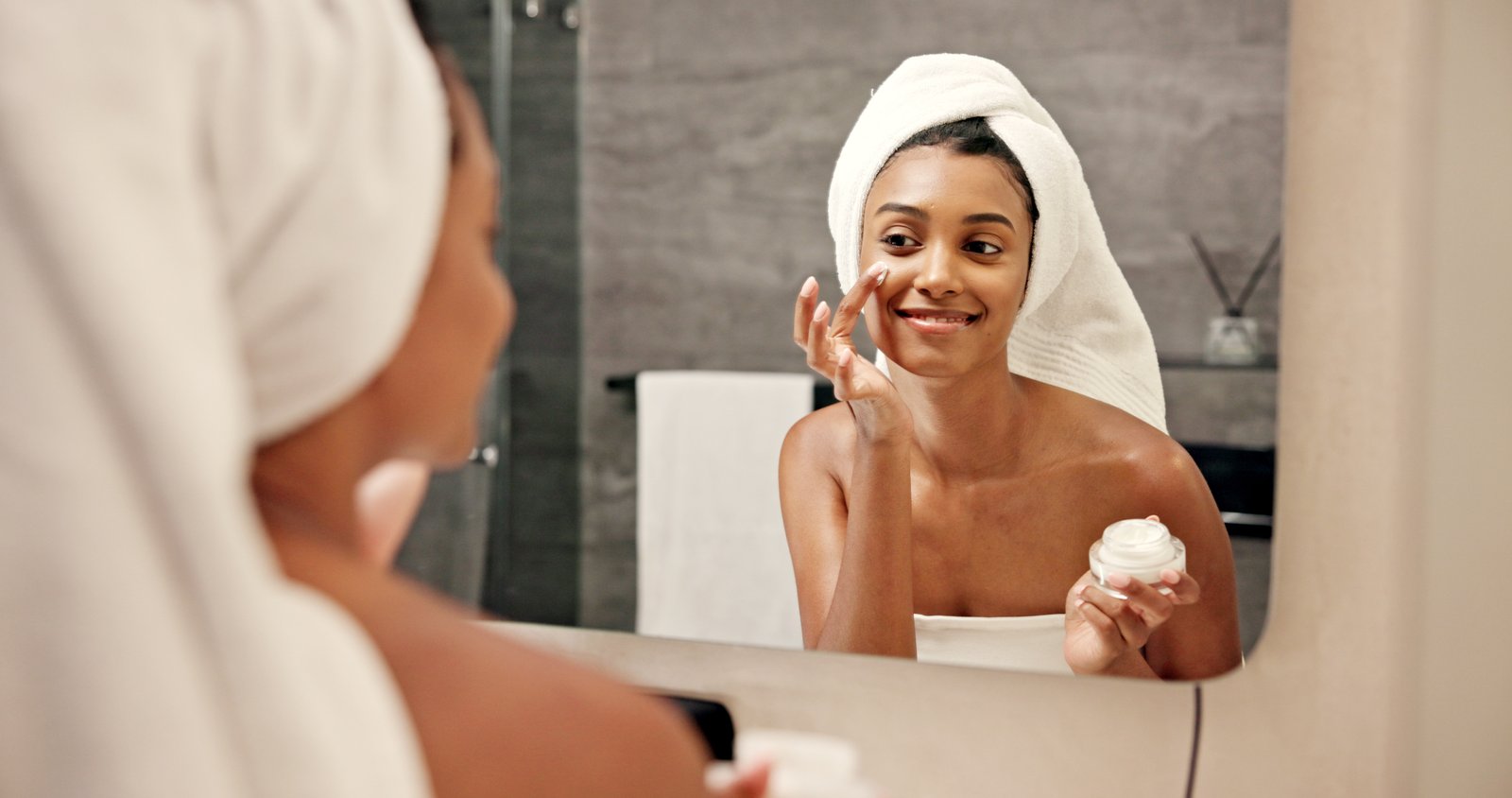 Woman with a towel wrapped on her head applying face cream in front of a bathroom mirror