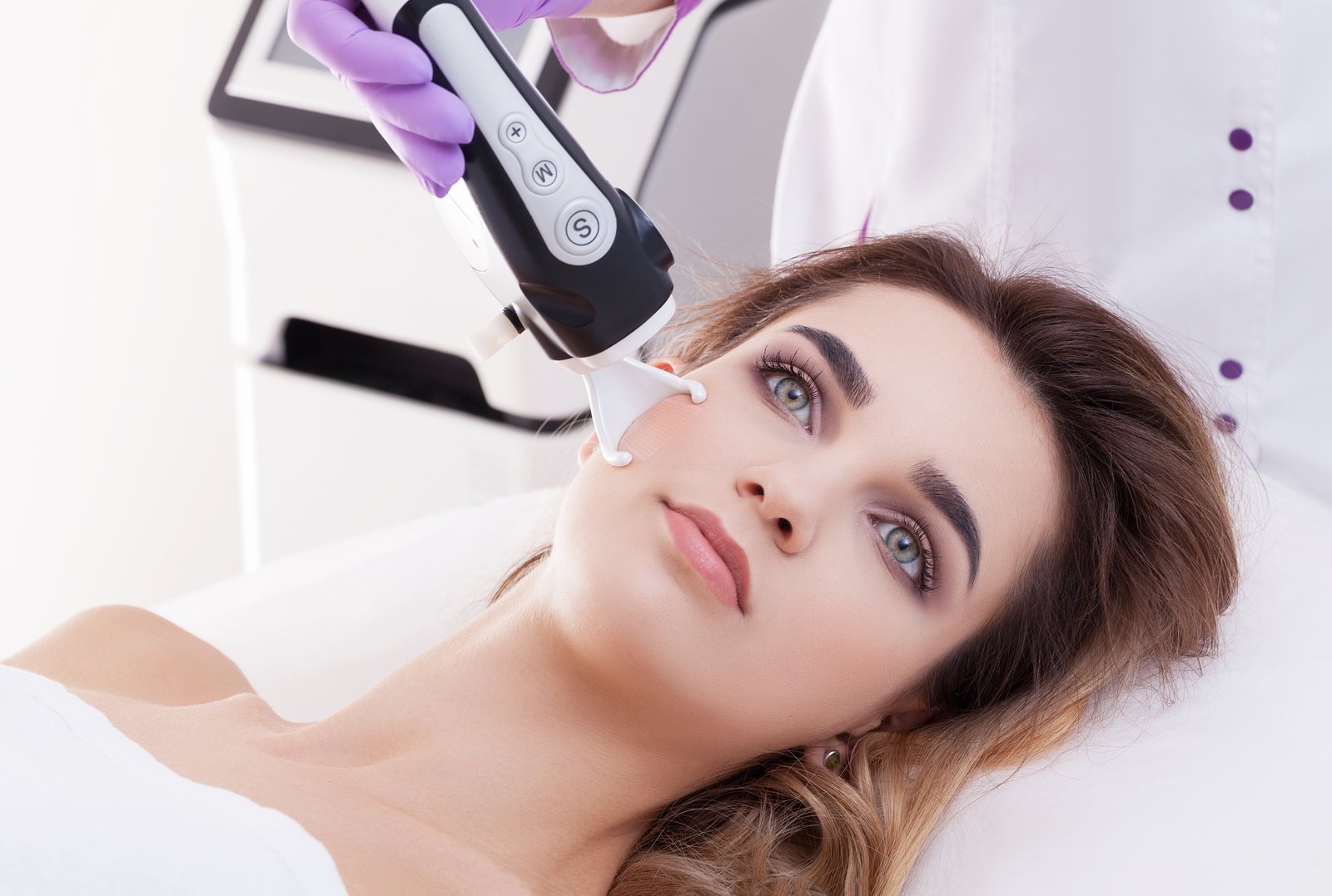 Patient reclining in a clinic chair while a laser handpiece is passed over the cheek